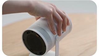 A hand is pushing the Freestyle downwards to project straight. The background then changes to show The Freestyle in a dark bedroom projecting an image to the wall then projecting upwards to the ceiling.